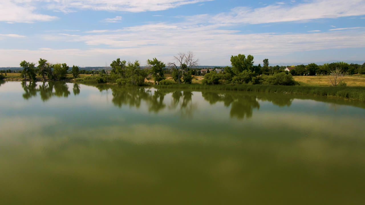 Broomfield, Colorado lake view with still green water, soft cloud reflections, a low grassy shoreline, clusters of trees, distant houses, and no visible people.