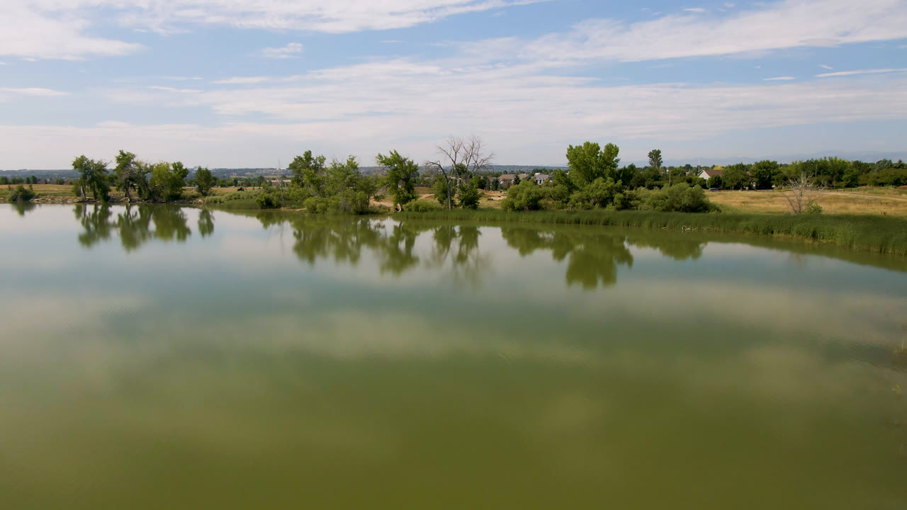 Broomfield, Colorado lake scene one second in, showing a broad green water foreground, reflected clouds, leafy trees along the shore, distant buildings, and no visible people.