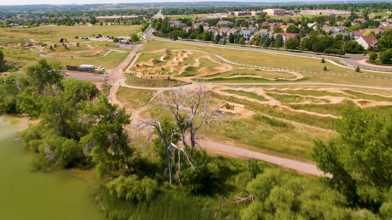 Aerial view of dirt track, pond edge, trees, roads, and suburban homes.