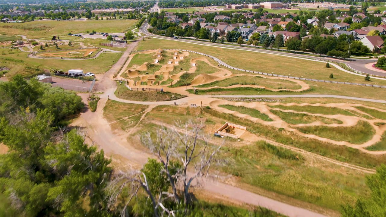 Wide aerial in Broomfield, Colorado with winding tan dirt track loops across dry grass, paved park paths and roads beyond, and no visible people.
