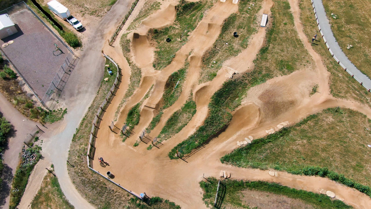 Overhead frame of a tan dirt bike track in the Denver United States collection, with paved paths, fencing, a small paved court, parked vehicles, and a tiny rider near the lower-left edge.