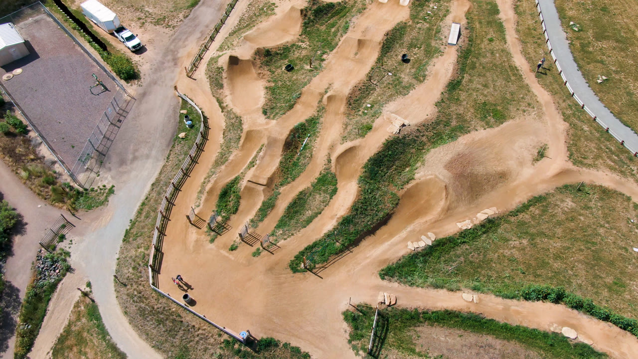 Aerial frame from the Denver United States clip showing the winding dirt course, grassy islands, roadside pavement, fencing, and a small rider with a bike near the lower-left trail bend.