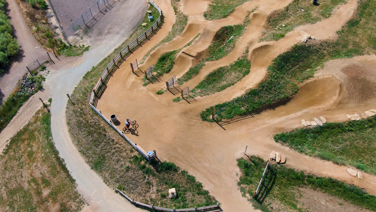 High overhead frame of a Broomfield, Colorado dirt BMX track with tan berms, grass strips, fencing, a paved path, and a distant rider near the lower-left junction.