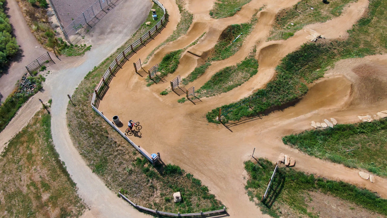 Overhead Broomfield, Colorado frame showing the BMX rider still small near the lower-left course entrance, with winding dirt routes and grassy banks across the frame.