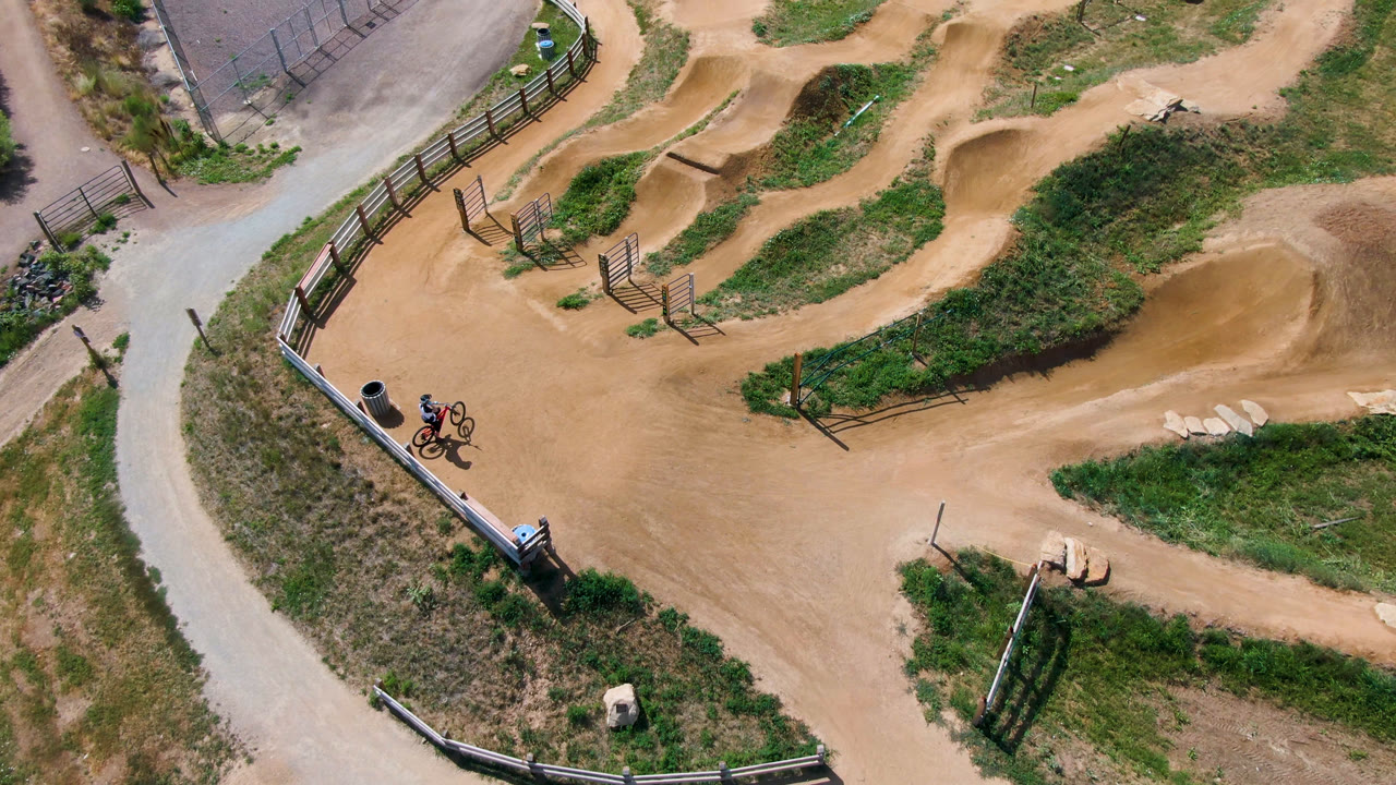 Overhead view of a cyclist paused beside a dirt bike track with fences and grass.