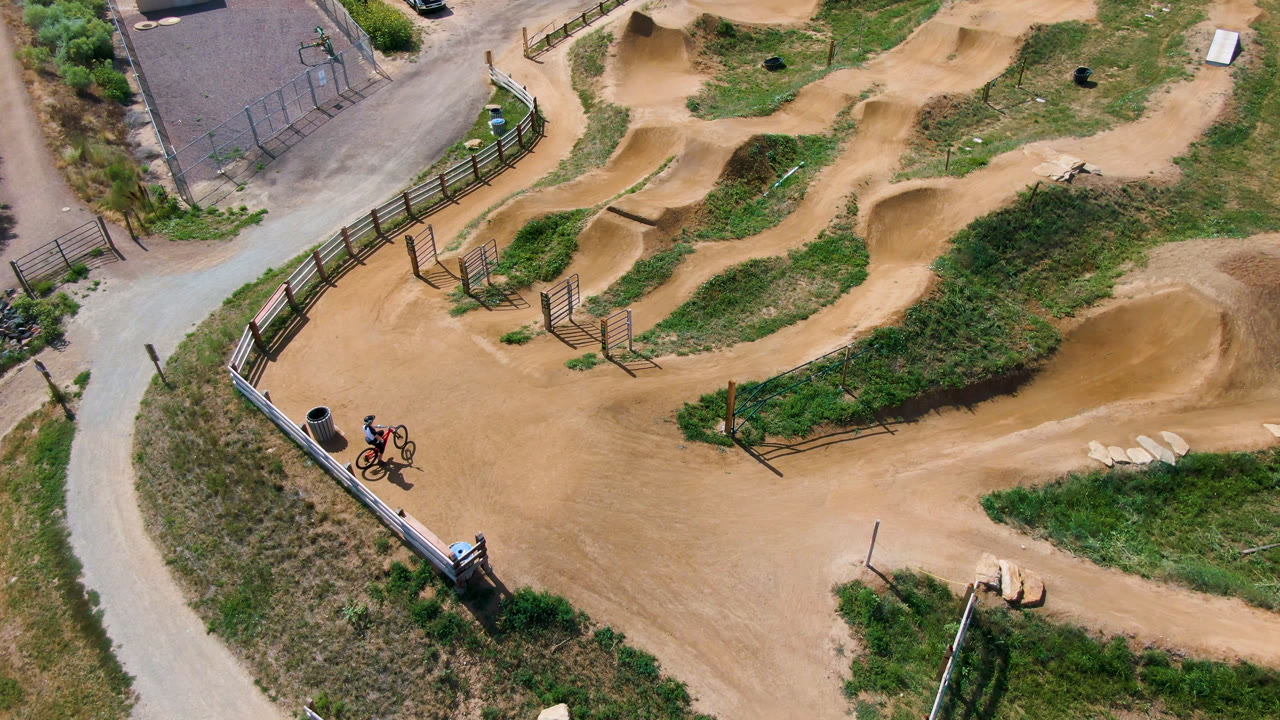 Opening overhead frame of a Broomfield, Colorado dirt bike course with winding tan paths, grass patches, fences, and two small riders near the lower-left edge.