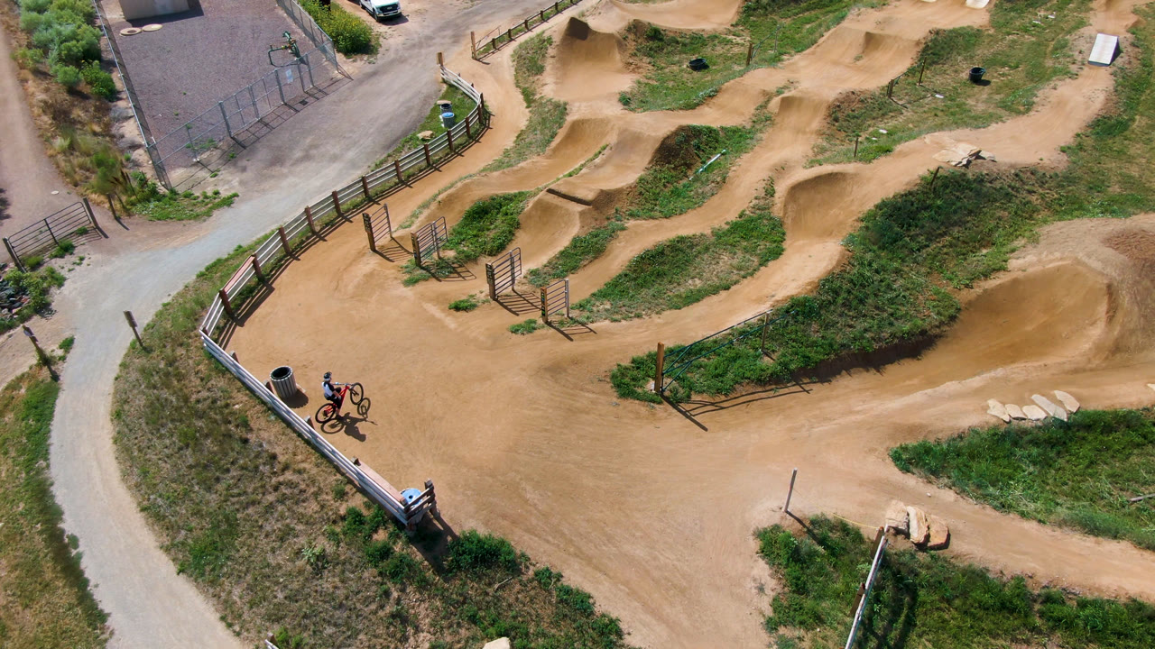 Drone-style overhead of a dirt bike park with tan jumps, green grass, fencing, and riders at the edge.