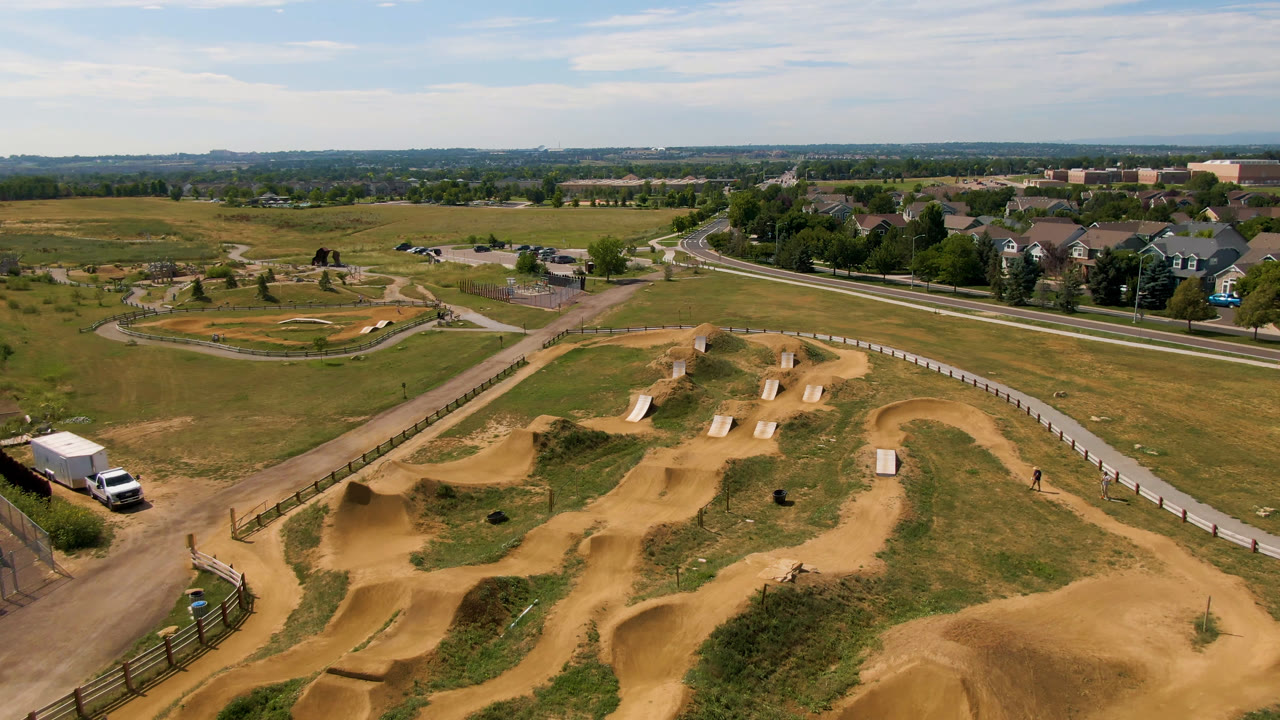 Aerial frame from Broomfield, Colorado, showing tan dirt track berms, a fence line, a white truck at lower left, open fields, distant homes, and no visible people.
