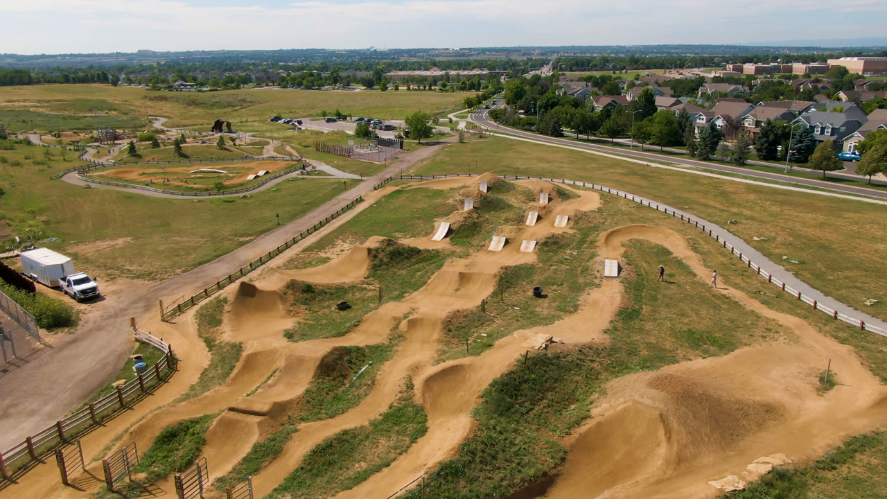 Drone wide frame over the Broomfield dirt bike park, with berms and jumps in the foreground, a road and grassy field beyond, suburban houses, and no visible people.