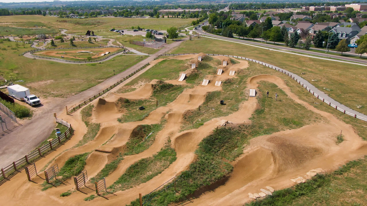 High aerial view in Broomfield, Colorado showing tan dirt bike jumps across grassy open land, a road and white vehicle at left, distant houses, and no clearly visible people.