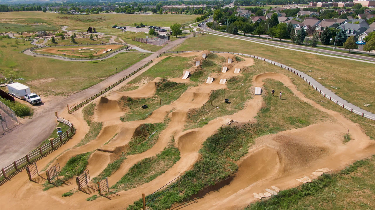 Wide drone-height view of dirt jumps and looping tracks with fields, fencing, and suburban houses beyond.