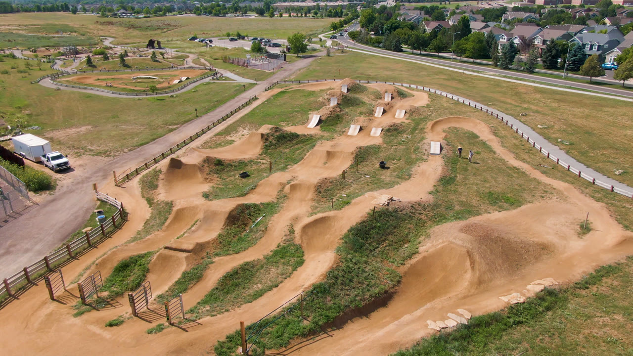 Wide Broomfield aerial showing a tan dirt course with berms and jumps in the foreground, roads and fields beyond, distant houses, and no clearly visible people.
