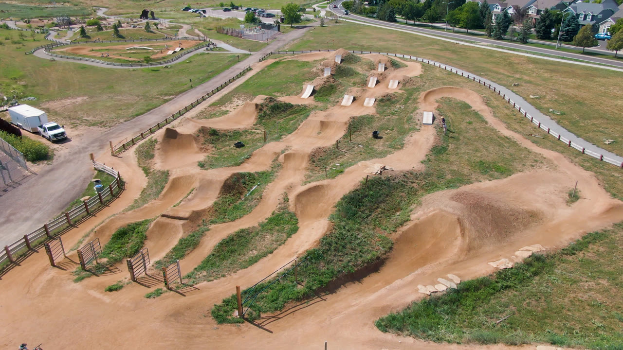 High-angle aerial frame in Broomfield, Colorado showing a tan dirt bike track with berms, grassy areas, fencing, a nearby road, and distant houses.