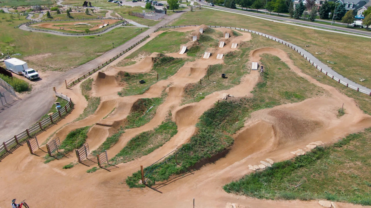 Second aerial frame in Broomfield, Colorado showing the dirt track from above, with winding tan paths, green grass, fencing, a white vehicle, road, and homes near the top edge.