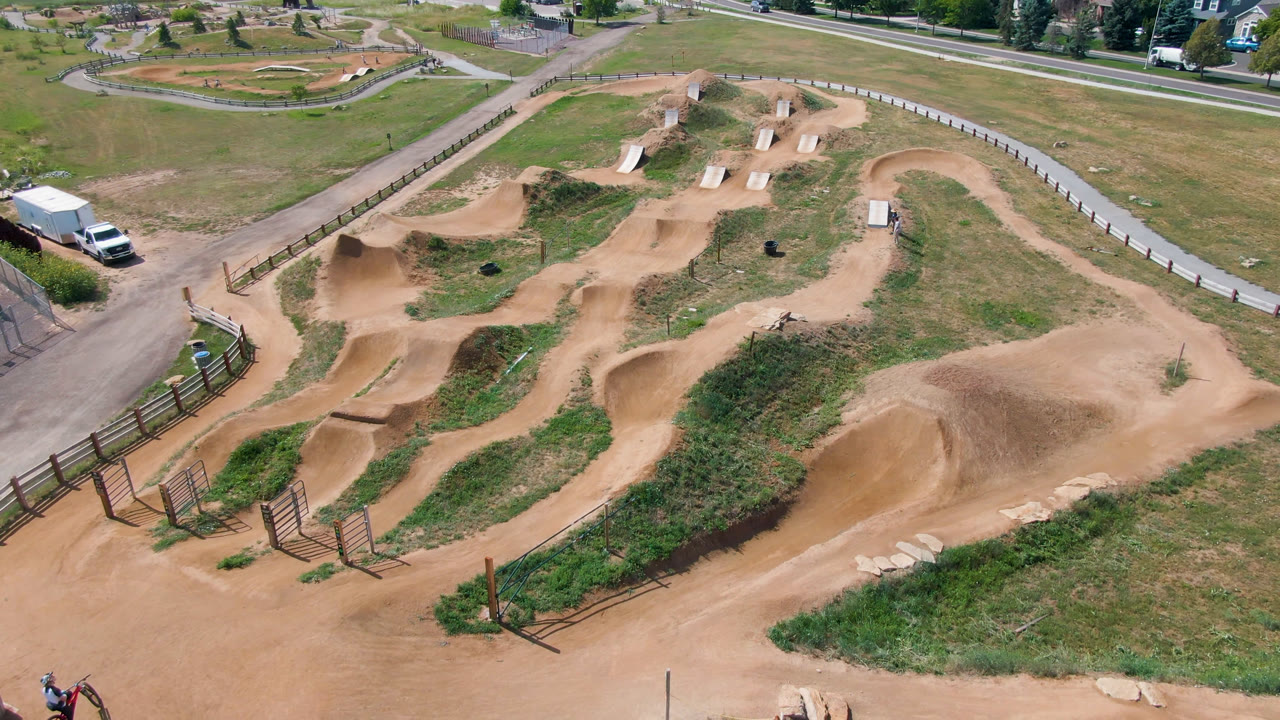 Wide aerial view of dirt bike park mounds and paths beside grass and a curved road.