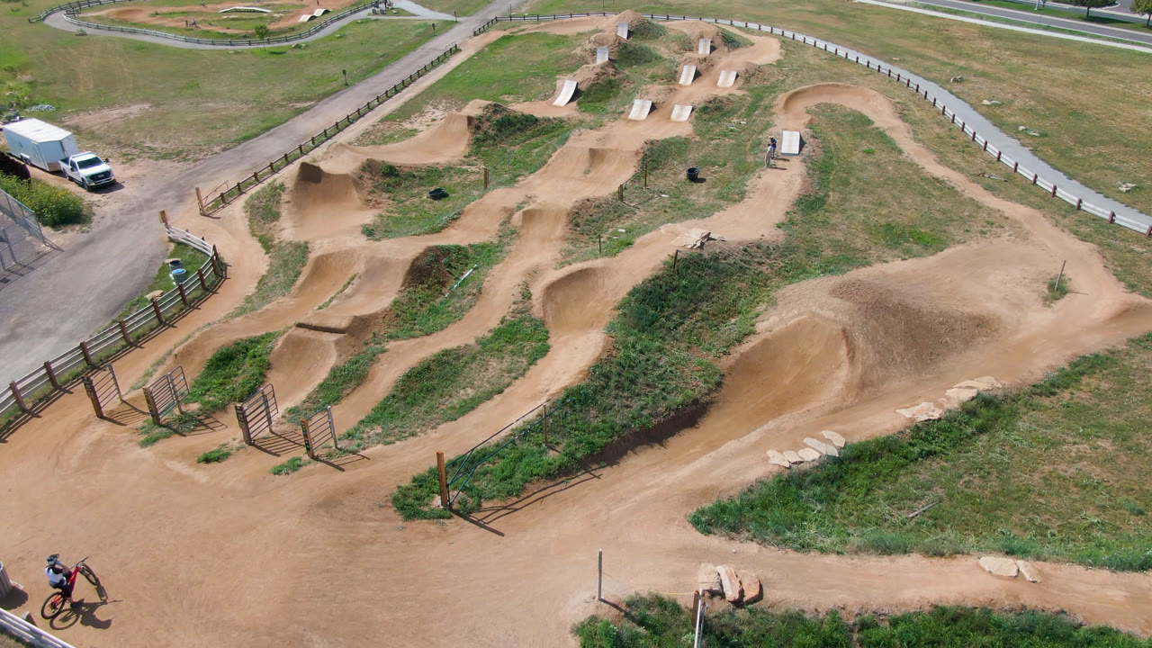 Overhead view in Broomfield, Colorado showing a tan dirt bike track, grass, fencing, parked vehicles, and small riders entering the lower edge of the frame.