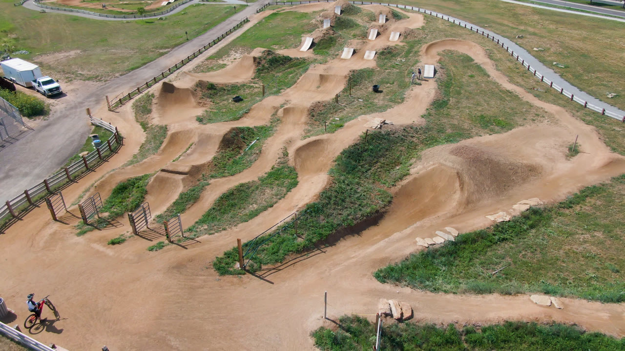 Aerial frame from Broomfield, Colorado with the winding dirt track centered, a cyclist near the bottom right, wood rails, grass patches, and parked vehicles at the upper left.