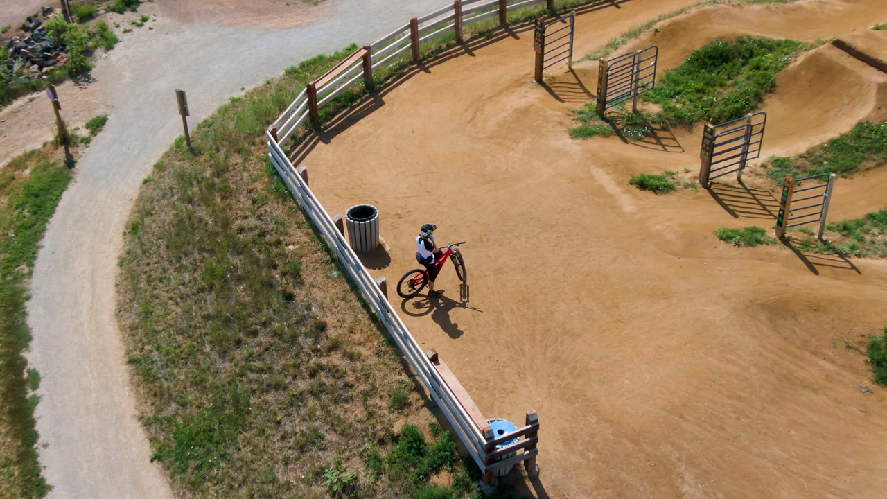 Overhead Denver, United States frame with the rider still near the lower-left fence line, a red bike, curved dirt berms, grass patches, a gravel road, and gate structures.