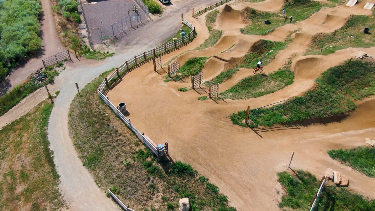 Overhead frame of a dirt bike park in Broomfield, Colorado, with tan track curves, fence lines, paved paths, grass, and a small distant bike or rider near the upper track.