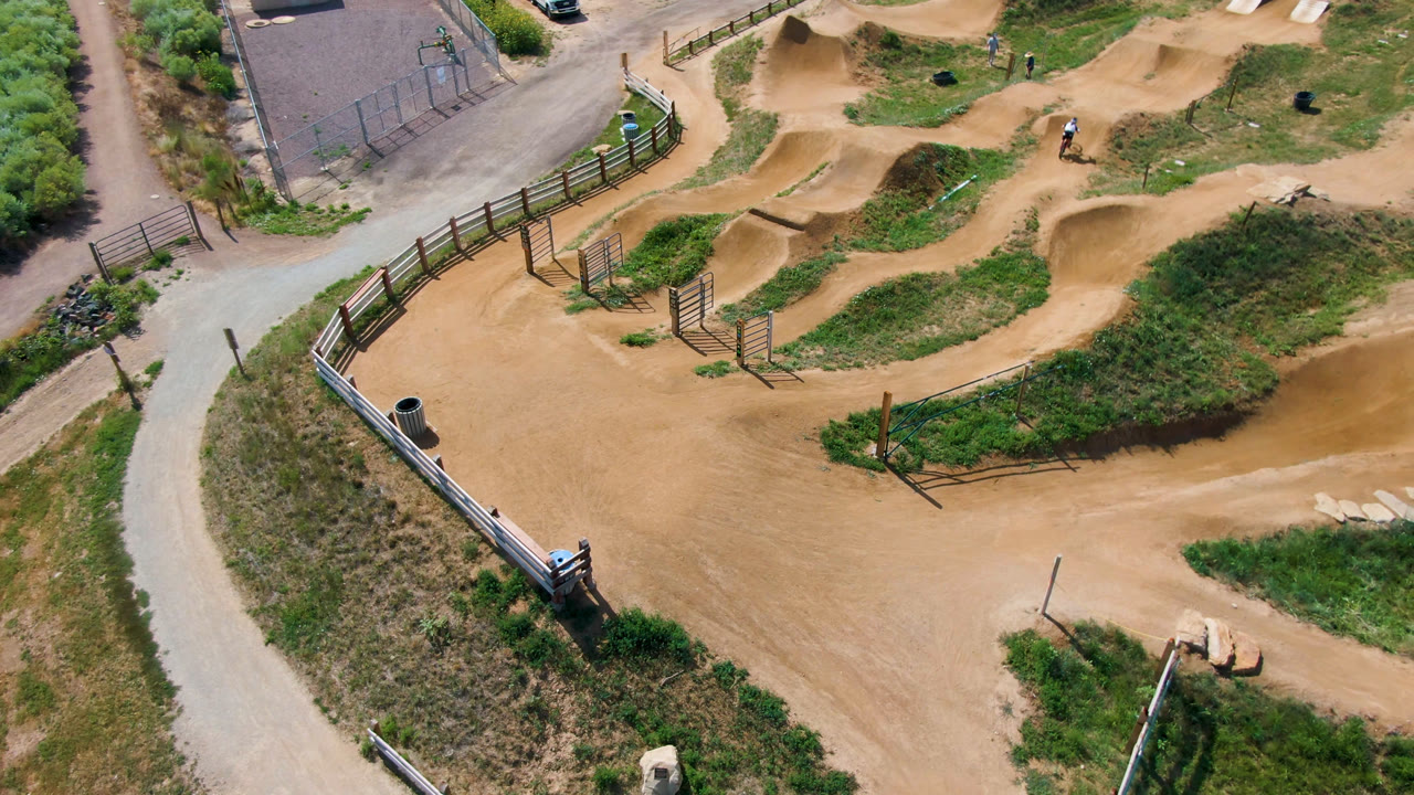 High overhead frame of the Broomfield dirt bike park with tan mounds, green borders, fence segments, path curves, and a tiny bike or rider near the upper right track.