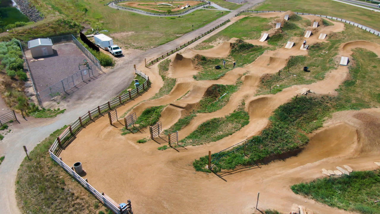 Overhead aerial frame of a Broomfield, Colorado dirt bike park with winding tan tracks, grassy infields, a fenced entry, nearby pavement, and no visible people.