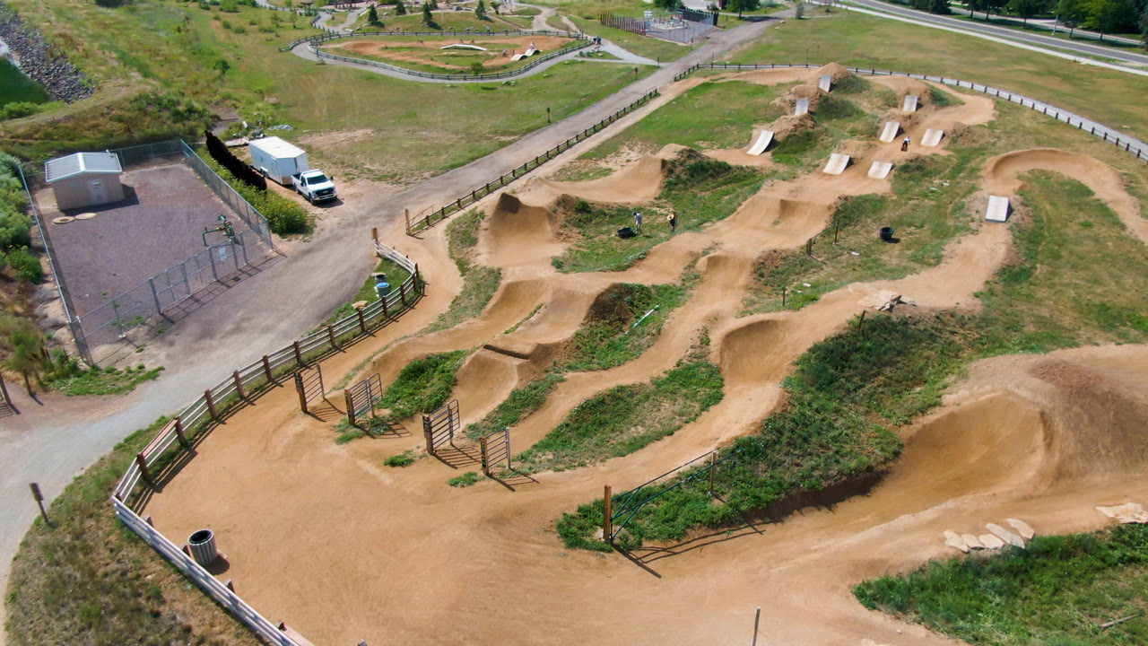 Second aerial frame over the Broomfield dirt bike park, showing the central dirt course, several ramp features, a roadside building, parked vehicles, and no visible people.