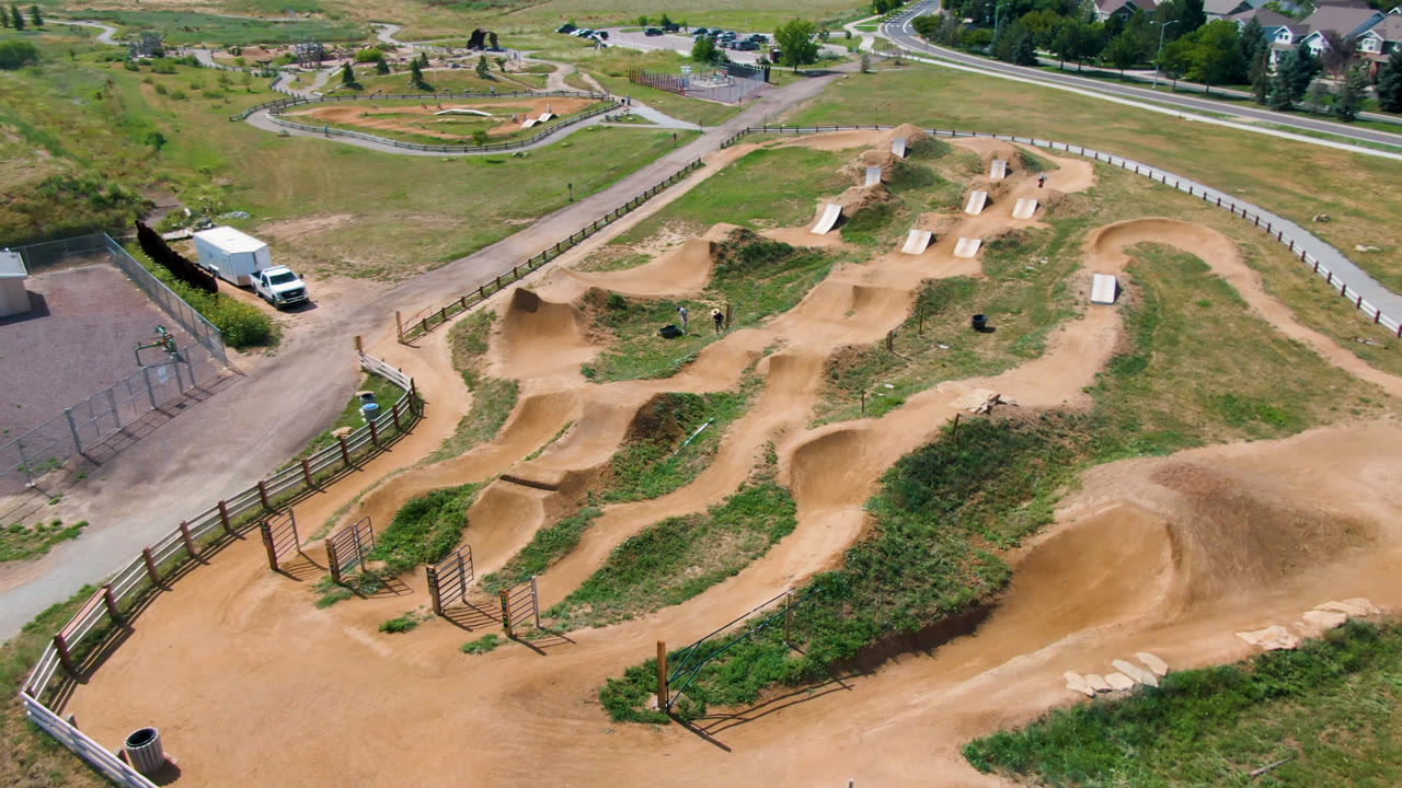 Wide aerial view of tan dirt bike paths, grassy areas, fences, roads, buildings, and houses in the distance.