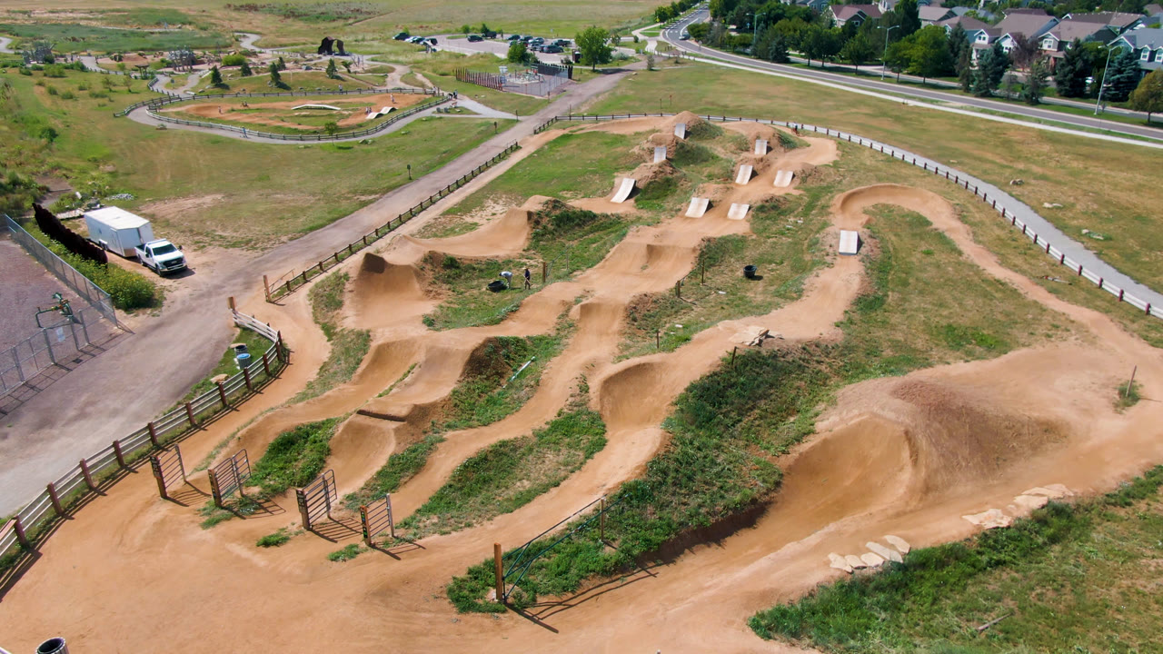 High aerial frame over a fenced dirt track in Broomfield, Colorado, with tan berms, green grass, nearby roads, distant houses, and no visible people.