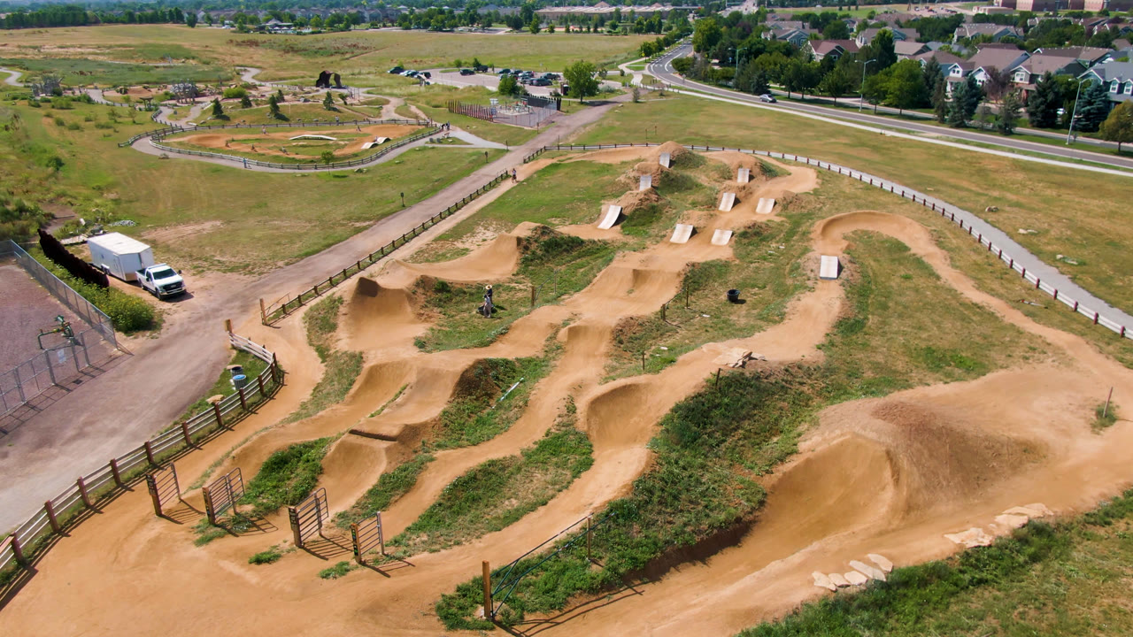 Wide drone view of dirt jumps, fencing, grass, and a nearby skate park.