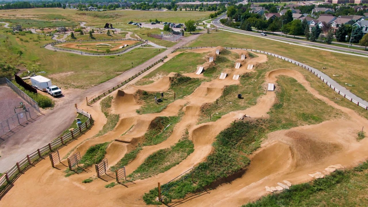 Third aerial frame over Broomfield, Colorado, with the larger foreground dirt mound, connected track lanes, skate park structures, roads, grassy open space, distant homes, and no visible people.