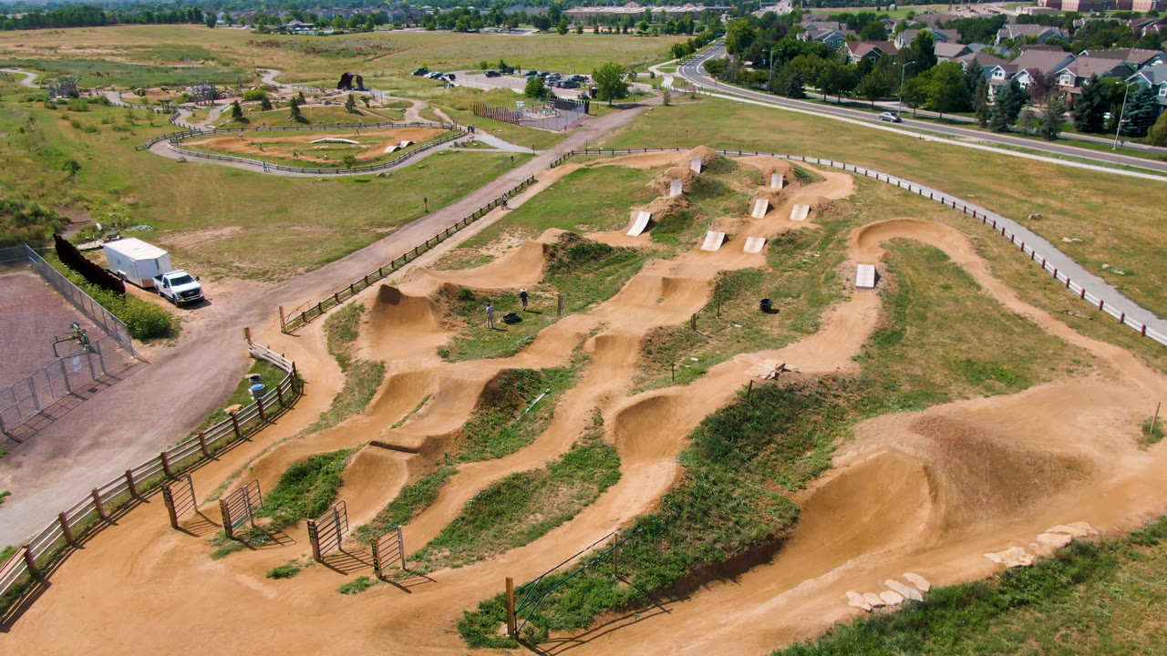 Aerial frame of an empty dirt track with skate park ramps in the background.