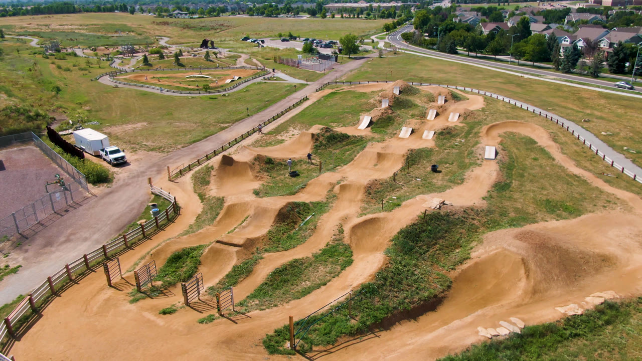 Opening aerial frame over a Broomfield, Colorado dirt skate and bike course, with tan jumps, fence lines, grass, roads, distant houses, and no visible people.