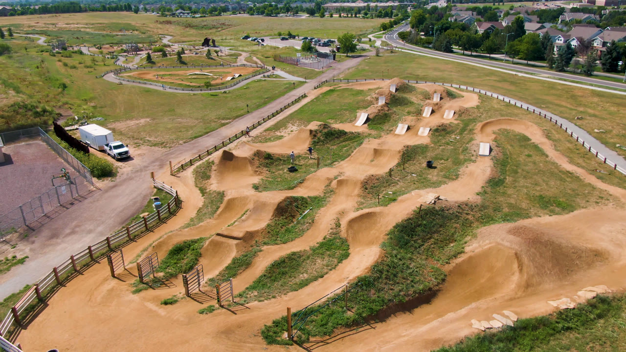 Wide aerial view of tan dirt jumps, fenced paths, grass, a white truck, and nearby roads.