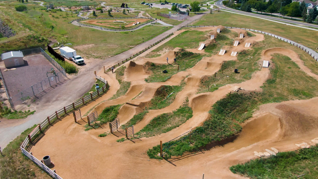 Opening overhead frame of a tan dirt course and fenced recreation area in Broomfield, Colorado, with grass, roads, a white vehicle, and no visible people.