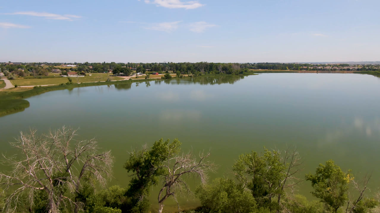 Wide aerial view in Broomfield, Colorado, with a green-blue lake, treetops at the bottom edge, distant shoreline, scattered clouds, and no visible people.