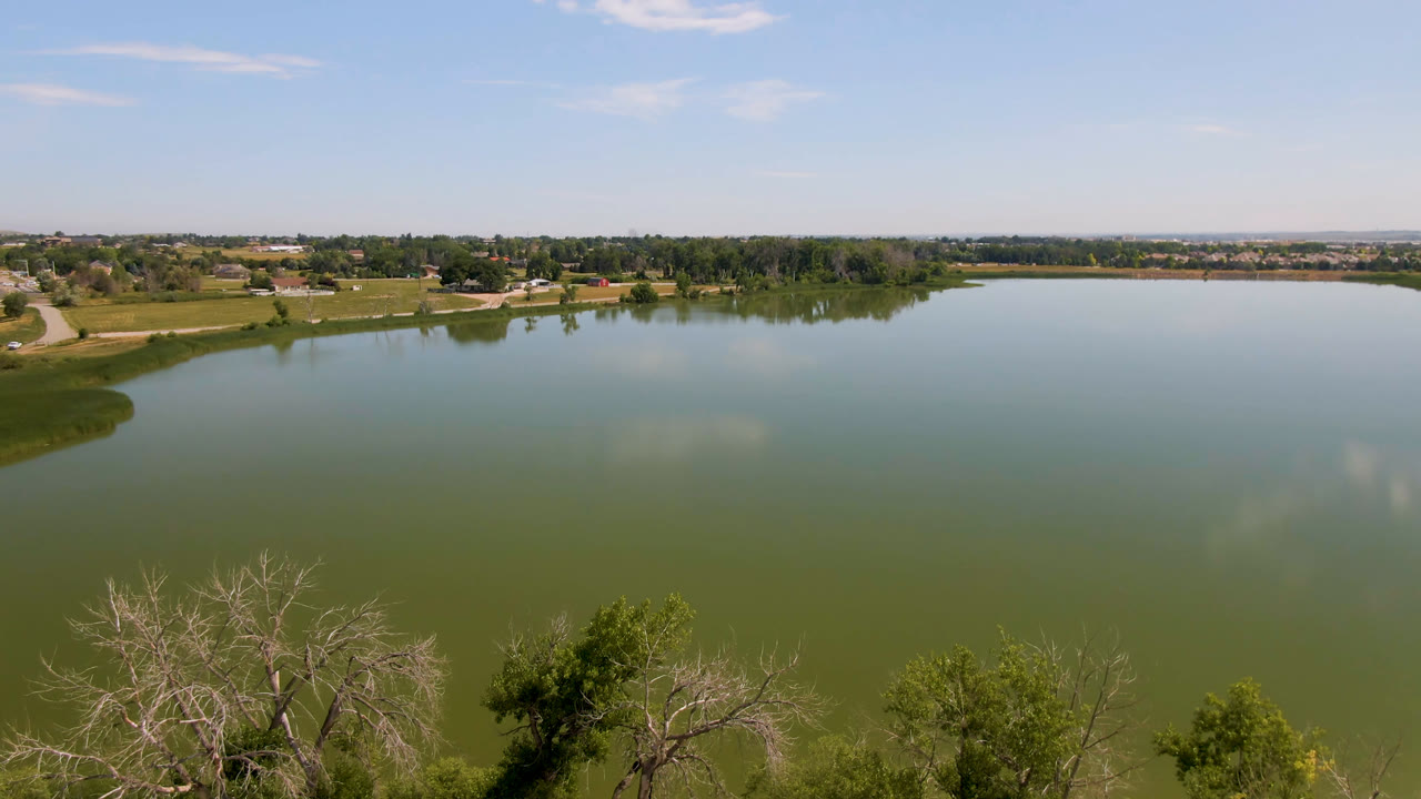 A broad Broomfield, Colorado lake surface fills the frame, with treetops and pale bare branches below, a distant tree line, fields, and no visible people.