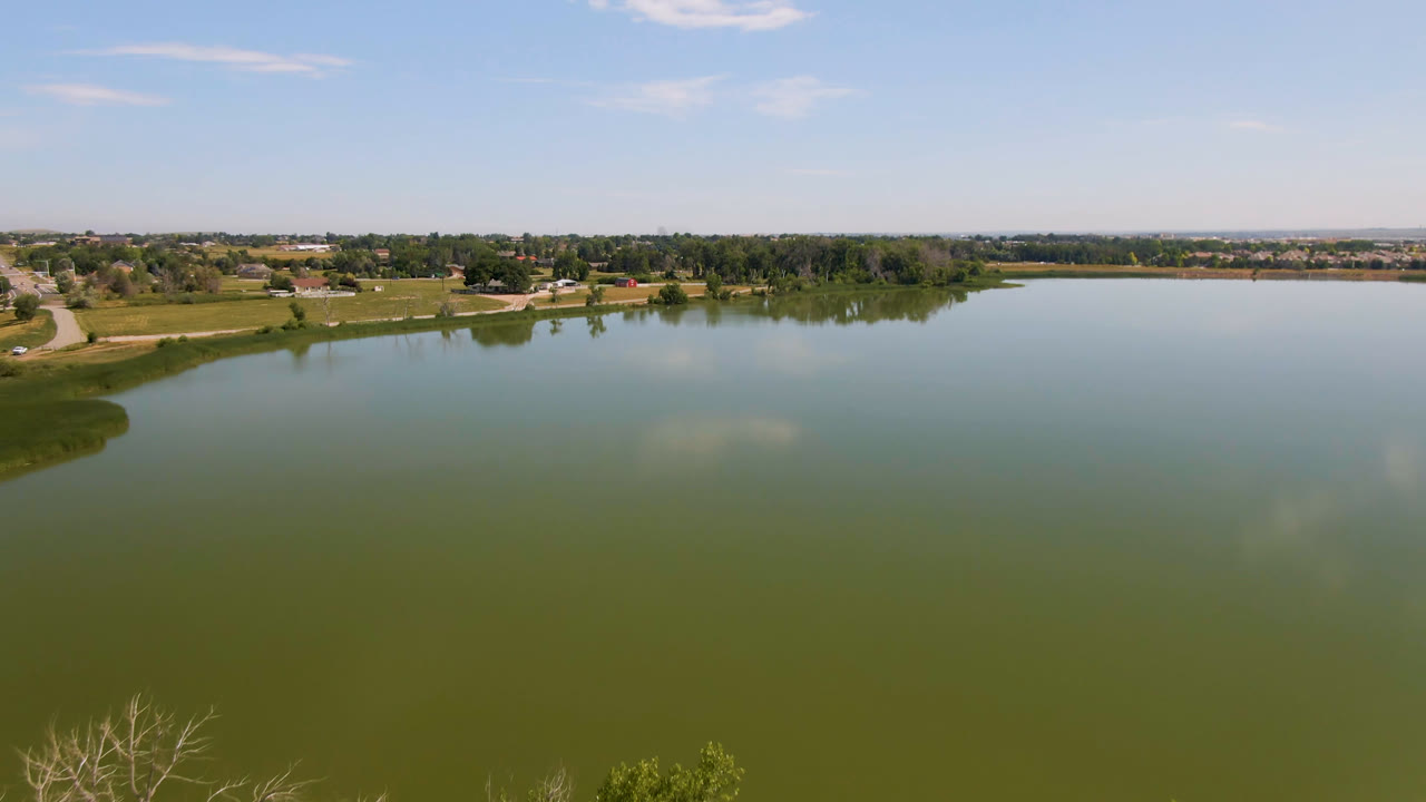 Aerial view over a broad green-blue lake in Broomfield, Colorado, with grassy shoreline at left, trees and low buildings across the horizon, and no visible people.