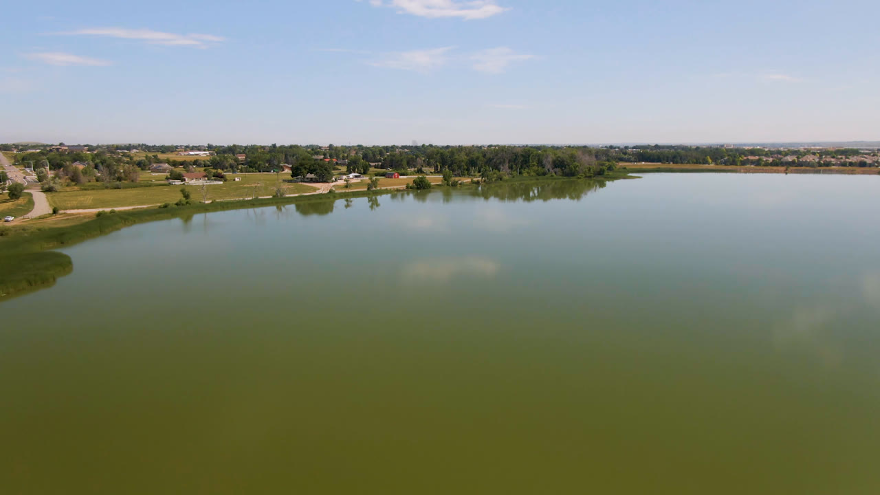 Large body of water with a small landform near the shore, trees, roads, and distant buildings.