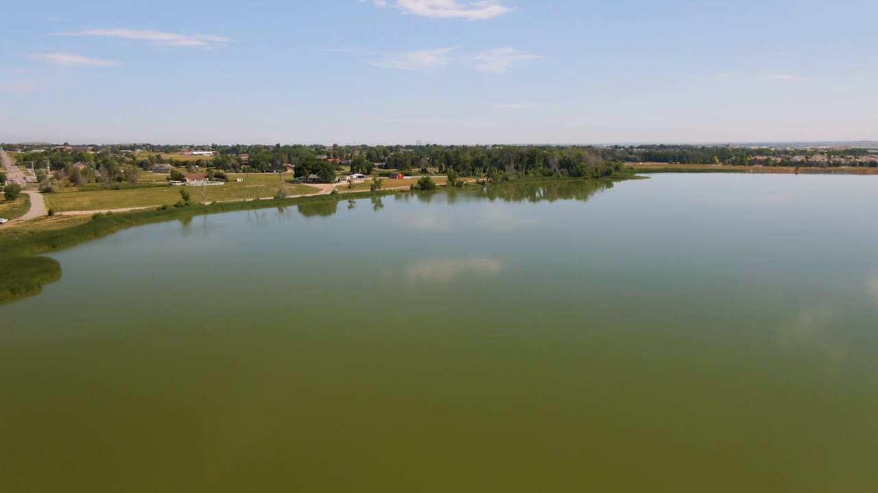 Broomfield, Colorado lake aerial with broad green water, a curved grassy edge on the left, trees and a road along the far shore, distant buildings, and no visible people.