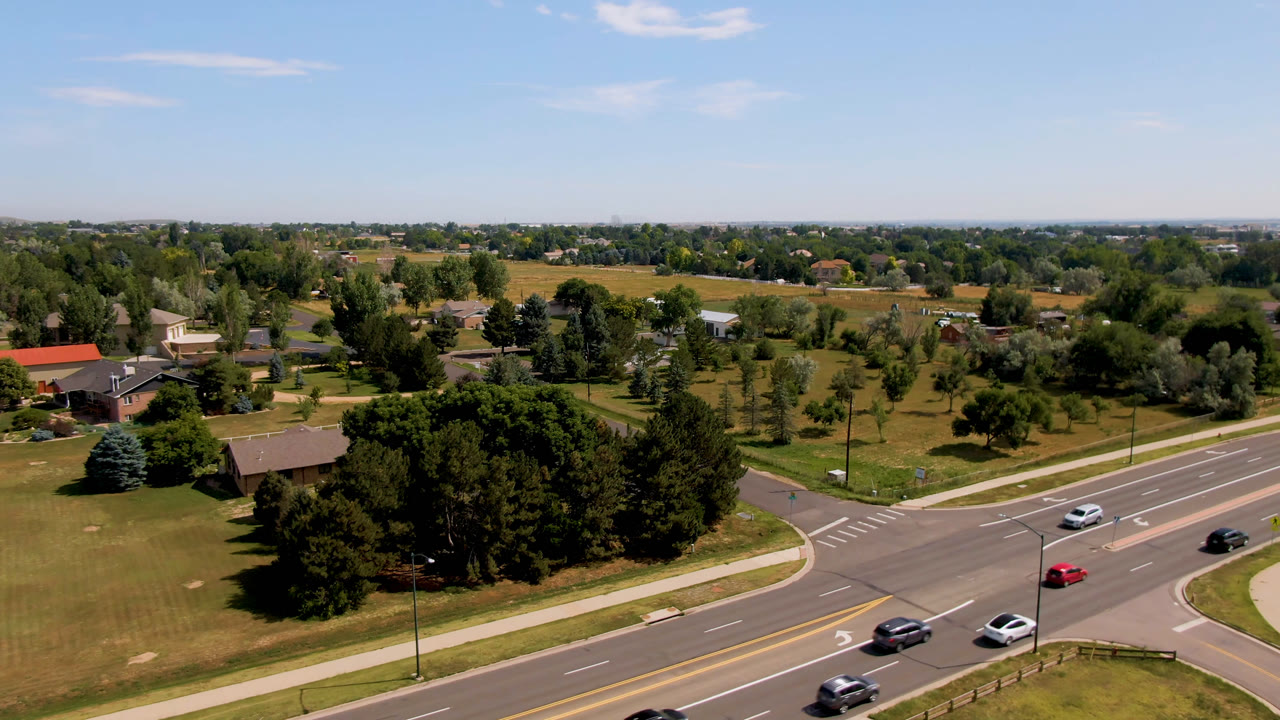 Aerial frame from Broomfield, Colorado showing cars on a multi-lane road beside lawns, dense trees, scattered buildings, open fields, blue sky, and no visible people.