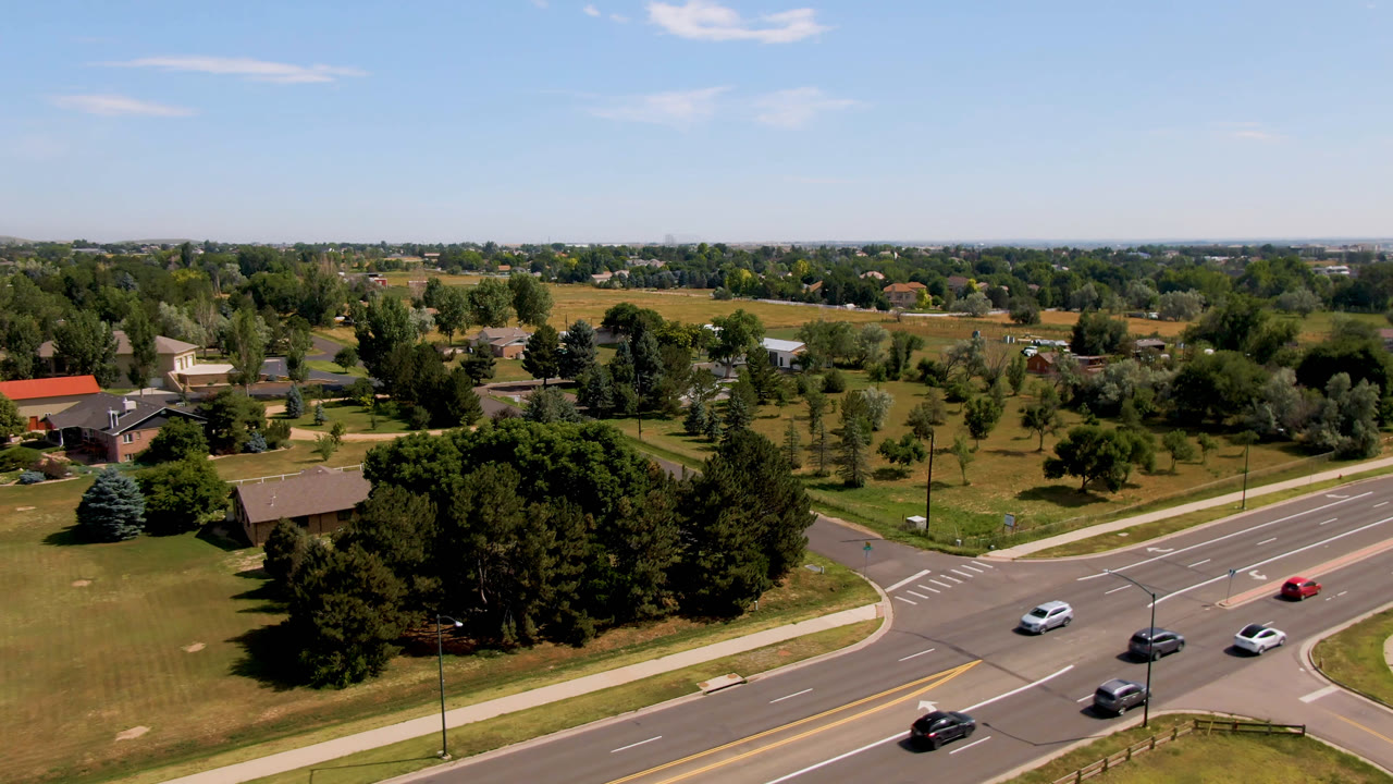 Second Broomfield, Colorado aerial frame with cars farther along the road, a crosswalk at the intersection, tree clusters, open grass, low buildings, and no visible people.