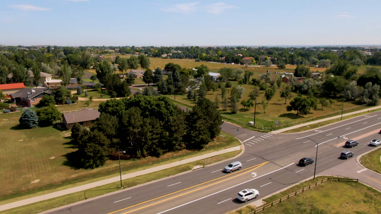 Third Broomfield, Colorado aerial frame showing vehicles on the foreground road, a broad tree mass, lawns, sidewalks, scattered buildings, distant fields, and no visible people.