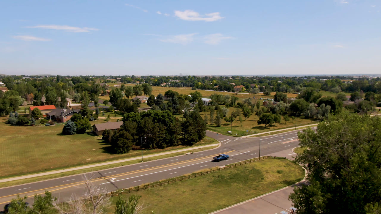 Aerial frame over Broomfield, Colorado showing a blue car on a multi-lane road beside grass, trees, scattered homes, and no visible people.