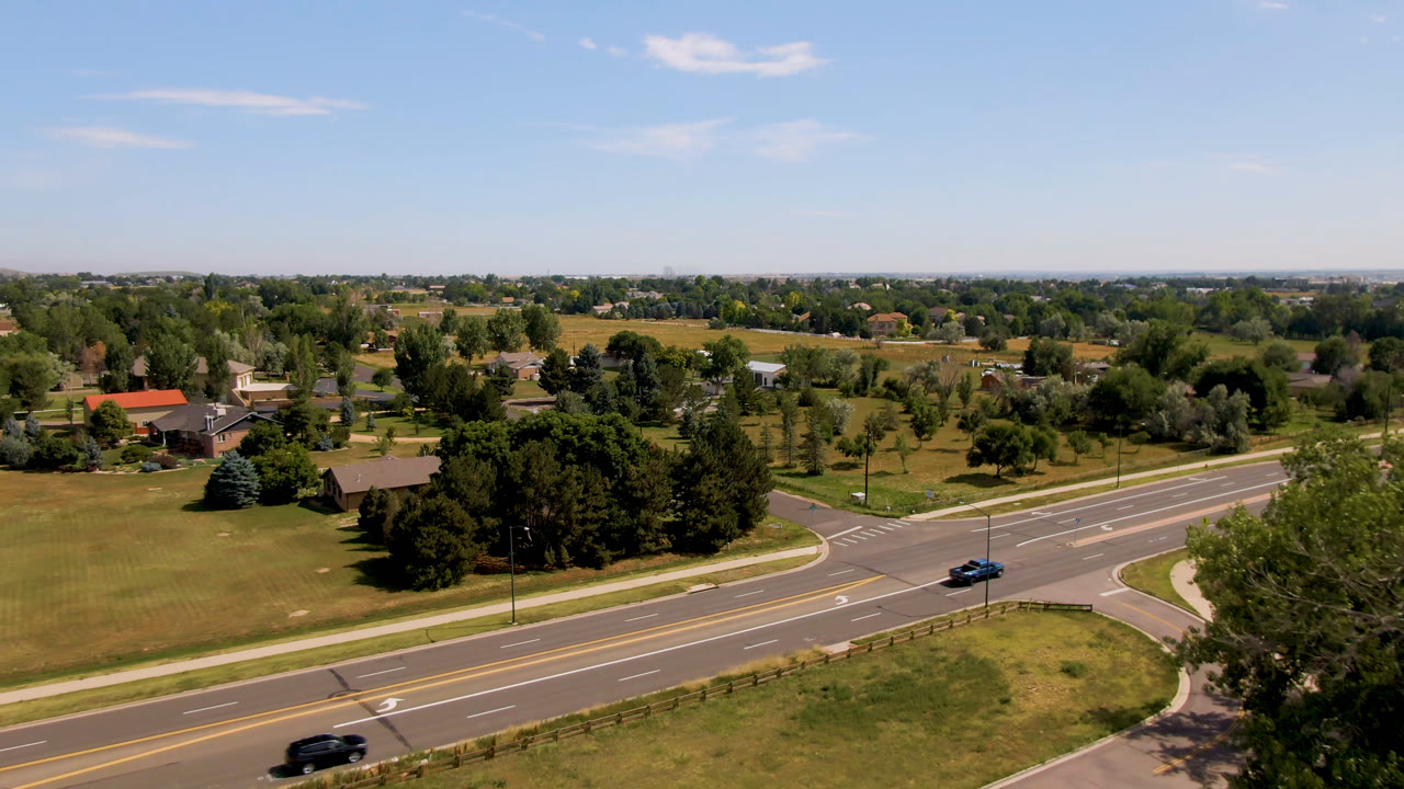 Aerial frame in Broomfield, Colorado with a blue car farther along the road, a dark vehicle at the lower edge, green lawns, trees, distant homes, and no visible people.