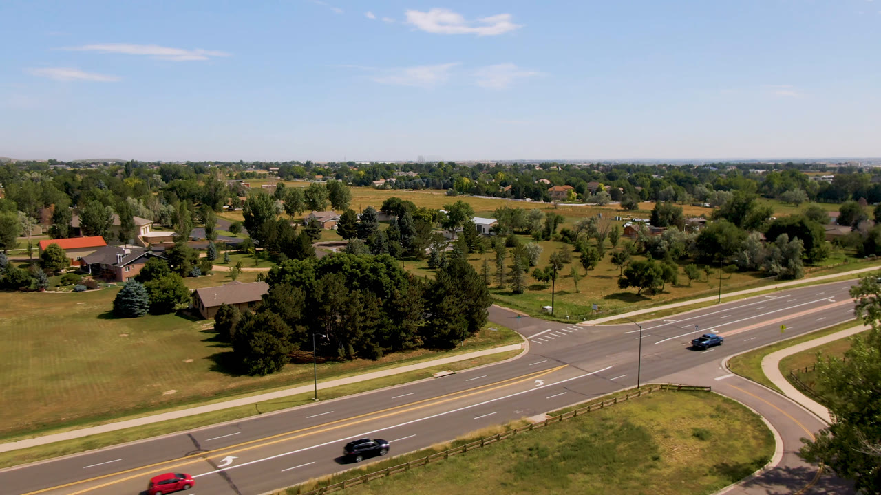 Aerial frame over Broomfield, Colorado showing a dark vehicle and red car on the road, with a curved intersection, lawns, tree clusters, homes, and no visible people.