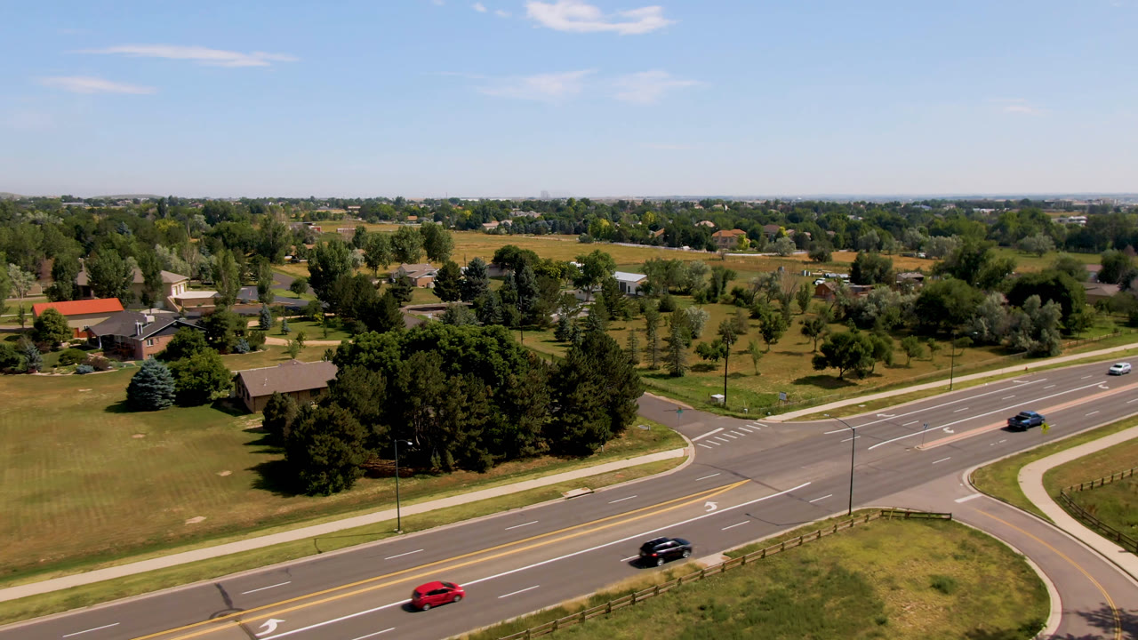 Wide aerial road view with red, dark, blue, and white cars near trees and homes.