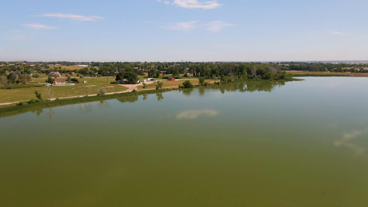Aerial view over green lake water toward fields, trees, and distant buildings.