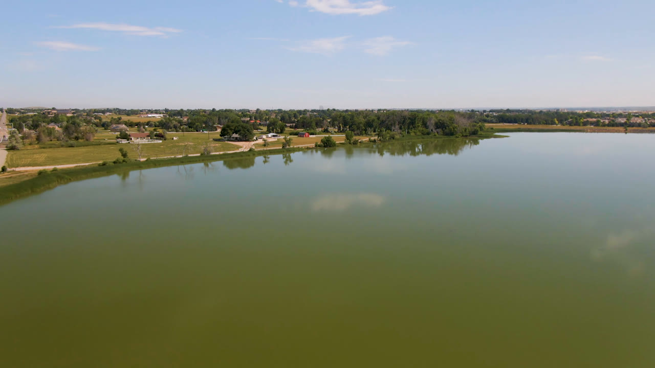Opening frame of a Broomfield, Colorado lake with green water in the foreground, a low shoreline with trees and houses beyond, scattered clouds overhead, and no visible people.