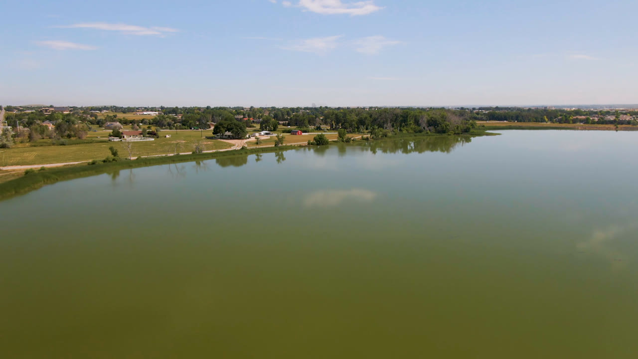 One second into the Broomfield, Colorado aerial view, the lake surface remains dominant with a distant band of trees, fields, small buildings, and no visible people.