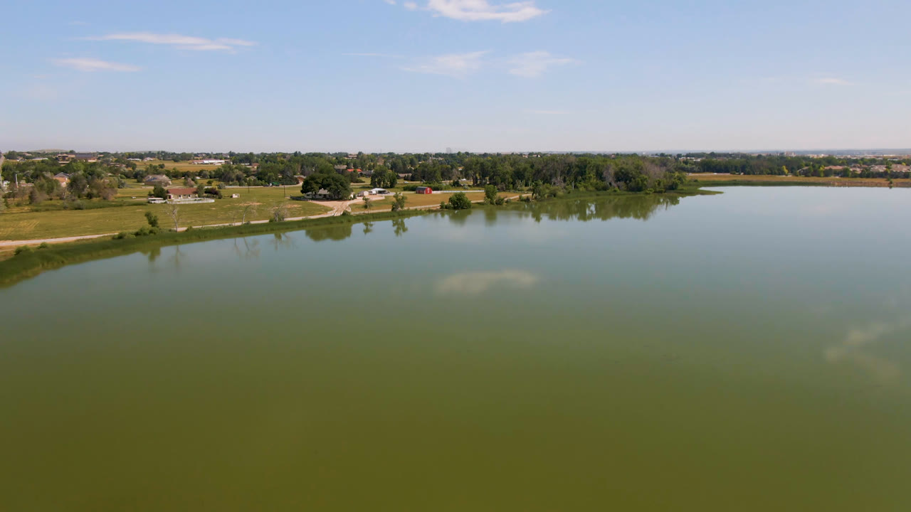 At 0:02, the Broomfield, Colorado lake fills most of the frame, with a thin horizon of trees, fields, and scattered houses under a bright sky, and no visible people.